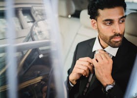 A bearded man in a black suit sitting inside a car, adjusting his tie and looking serious.