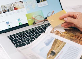 Hands holding credit card over laptop and magazine for online shopping and travel planning.